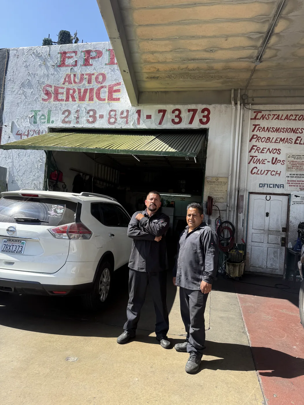 Two EPI Auto Service mechanics in work uniforms in front of the shop, with the painted EPI Auto Service sign and a vehicle in the bay behind them